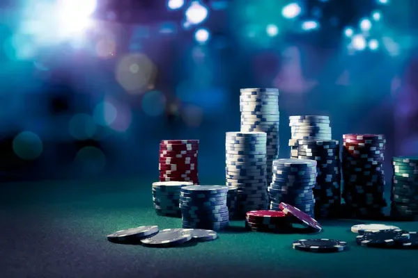 Pilas de fichas de casino blancas, rojas, negras y verdes sobre un tapete verde, con luces desenfocadas al fondo.